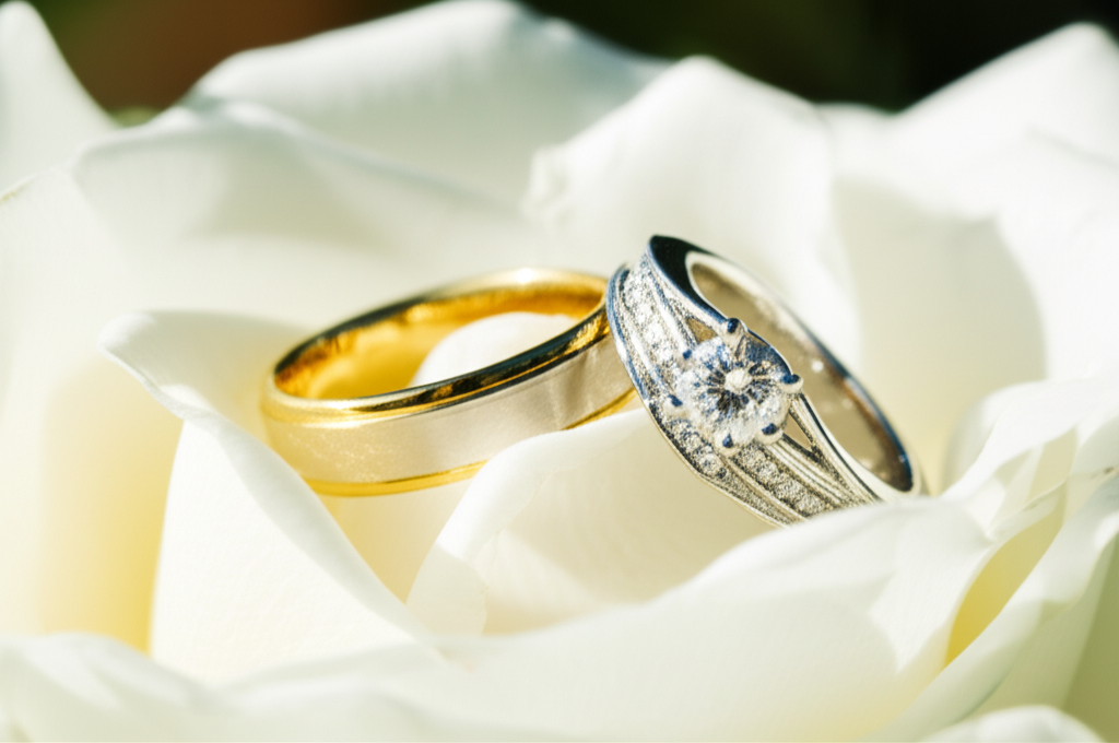So sánh nhẫn cưới kim cương và đá quý với so sánh nhẫn cưới 6 Ảnh chụp cận cảnh cặp nhẫn cưới, một chiếc nhẫn vàng trơn và một chiếc nhẫn bạch kim đính kim cương và đá quý, đặt trên những cánh hoa hồng trắng trong một khu vườn ngập nắng.